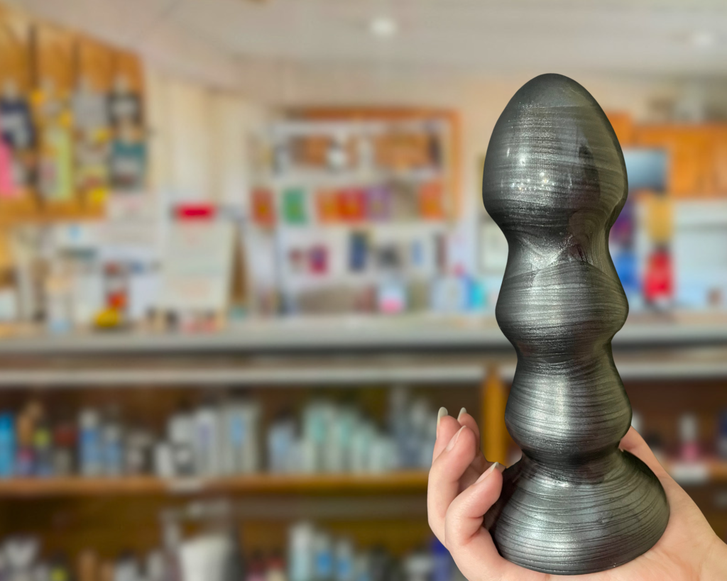 Hand holding a black and grey dildo with the sex store blurred as background